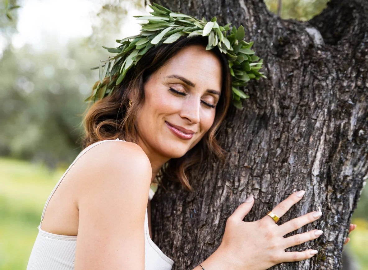 Woman wearing an olive-leaf crown, eyes closed, hugging an olive tree trunk